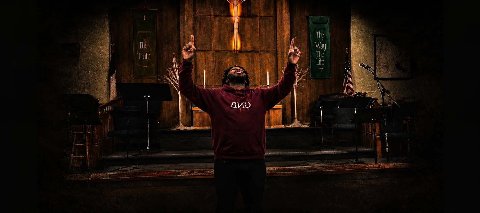 A worshipper in a burgundy GNP hoodie raises both hands toward heaven in the sanctuary